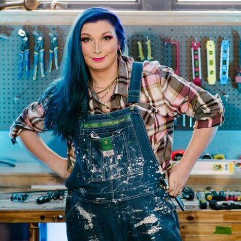 Mercury Stardust, who has purple hair and is wearing paint-splashed overalls over a purple dominant plaid shirt, stands with their hands on their hips and smiles towards the camera.