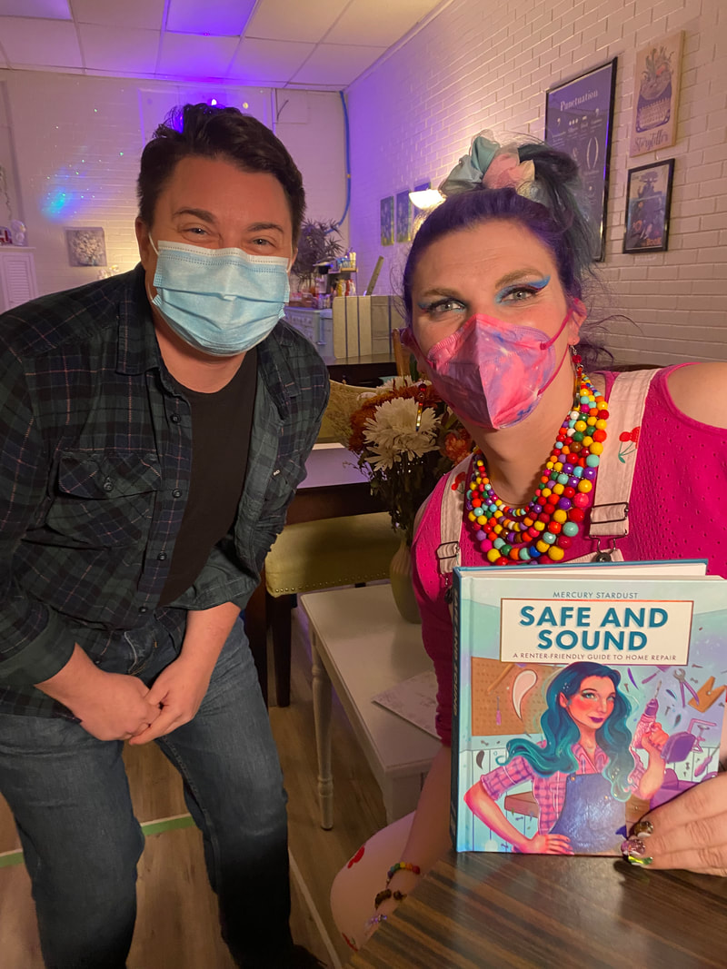 Derek, with short brown hair and wearing a green plaid shirt and denim jeans, crouches down to take a photo with with Mercury, who has purple hair and is wearing a hot pink shirt under light pink overalls with cherries as she holds up a copy of her book. Both are wearing face masks.